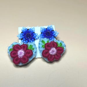 Puede incluir: Un par de pendientes hechos a mano con un diseño de flor azul con cuentas sobre una base en forma de corazón azul claro con una flor roja. Los pendientes están sujetos a una tarjeta blanca sobre un fondo neutro.