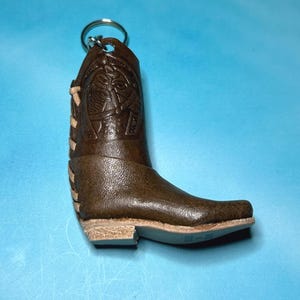 May include: A brown leather cowboy boot keychain with a silver keyring. The boot features decorative stitching and a textured surface. The heel and sole are a lighter tan color. The boot is a miniature replica of a classic Western boot.