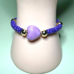 Puede incluir: Un brazalete con una cuenta en forma de corazón púrpura. El brazalete presenta una cuenta central en forma de corazón, cuentas doradas y una banda azul tejida. El brazalete se muestra sobre una superficie de color claro sobre un fondo verde claro.