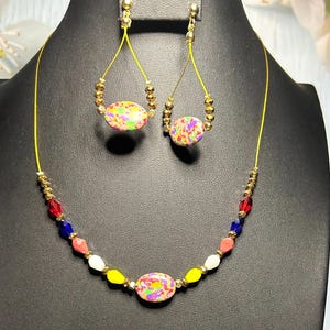 Puede incluir: Un conjunto de joyas colorido que incluye un collar y pendientes a juego. El collar presenta una cuenta ovalada central con un patrón moteado, complementada con cuentas más pequeñas de varios colores. Los pendientes reflejan el diseño de la cuenta central, suspendidos de alambres dorados.