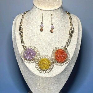 May include: A gold-toned necklace and earring set displayed on a mannequin. The necklace features three large, colorful floral designs in purple, yellow, and orange. The earrings have small, round, pinkish-purple beads. The jewelry is set against a blue background.