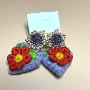 Puede incluir: Pendientes hechos a mano con la parte superior de cuentas en tonos morados y dorados, combinados con detalles de flores de fieltro. Los pendientes tienen un respaldo azul claro y están adornados con flores rojas y amarillas bordadas.