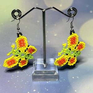 Puede incluir: Un par de pendientes de mariposa con cuentas. Los pendientes son amarillos, naranjas, verdes y negros. Las alas de mariposa tienen un patrón de cuentas detallado. Los pendientes se exhiben en un soporte de pendientes de metal plateado con una base transparente.