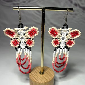 Puede incluir: Pendientes hechos a mano con cuentas con un diseño de mariposa. Los pendientes tienen una base blanca con detalles en rojo y hebras colgantes de cuentas rojas, rosas y blancas. Los pendientes se exhiben en un soporte dorado.