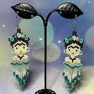 Puede incluir: Pendientes de cuentas hechos a mano con un diseño de retrato. Los pendientes presentan una cara color crema, cabello negro y boca roja. Tienen una borla de cuentas turquesa y blanca. Los pendientes se exhiben en un soporte negro.