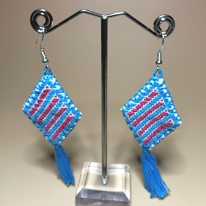 Puede incluir: Un par de pendientes hechos a mano con forma de diamante. Los pendientes son azules con rayas rosas y blancas y una borla azul. Los pendientes se exhiben en un soporte de metal plateado con una base transparente.