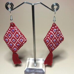 Puede incluir: Un par de pendientes rojos y blancos en forma de diamante con un patrón geométrico y pequeñas borlas rojas. Los pendientes se exhiben en un soporte plateado para pendientes sobre un fondo neutro.