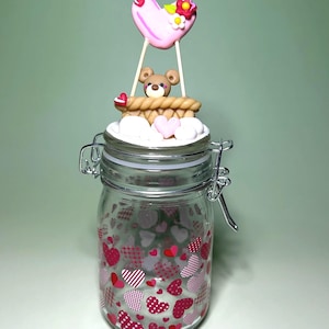 Könnte beinhalten: Ein klares Glas mit roten und rosa Herzen verziert. Das Glas hat einen Metallverschluss und einen Deckel mit einer dekorativen Szene. Die Szene zeigt einen Teddybären in einem Korb, ein rosa Herz und kleine Blumen, die ein romantisches Thema bilden.