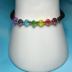 May include: A beaded bracelet with a rainbow of colors. The bracelet features faceted beads in shades of purple, blue, green, yellow, and red, separated by small gold-colored beads. The bracelet is displayed on a white surface against a blue background.
