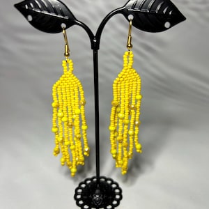 Puede incluir: Un par de pendientes de cuentas amarillas con un diseño de flecos. Los pendientes se exhiben en un soporte de pendientes de metal negro. Los pendientes tienen un gancho dorado.