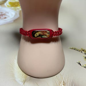 Könnte beinhalten: Rotes geflochtenes Armband mit einem rechteckigen Mittelstück mit einem religiösen Bild. Das Armband wird an einem Schaufensterpuppenarm präsentiert. Das Bild auf dem Mittelstück zeigt eine religiöse Figur.