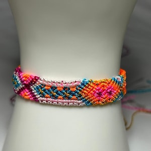 Puede incluir: Una colorida pulsera de la amistad con un patrón tejido. La pulsera presenta tonos vibrantes de rosa, naranja, azul y blanco. La pulsera mide aproximadamente 3,8 cm de ancho y se muestra sobre una superficie blanca.