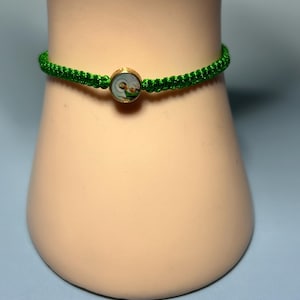 May include: A green macrame bracelet with a gold-colored circular charm. The charm features a small, detailed image. The bracelet is displayed on a light-colored, rounded surface against a blue background.