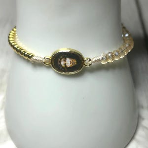 May include: Gold-toned bracelet featuring an oval pendant with a religious image. The bracelet has a beaded design with gold and clear crystal beads. The bracelet is displayed on a white surface.