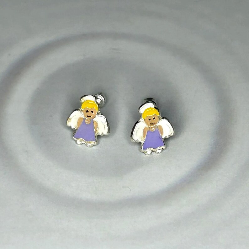 two small silver earrings with angel designs, placed on a gray surface.