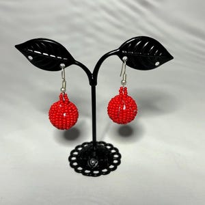 Puede incluir: Un par de pendientes de bola roja con cuentas, colgados de ganchos plateados. Los pendientes se exhiben en un soporte de metal negro con detalles en forma de hoja y una base decorativa. Los pendientes son de color rojo vibrante.
