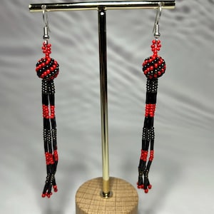 Puede incluir: Un par de pendientes de cuentas rojas y negras. Cada pendiente presenta una parte superior redonda con cuentas y un diseño largo y colgante. Los pendientes se exhiben en un soporte dorado con una base de madera.