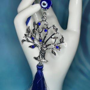 May include: A silver-toned tree of life charm with blue accents and a matching tassel. The charm features a blue evil eye bead and is suspended from a blue cord. The tree's leaves are adorned with blue gemstones.