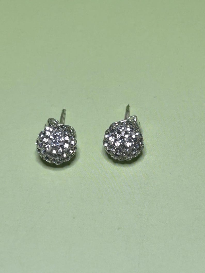A pair of silver earrings with small, round, sparkling stones on a light green background.