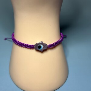 Könnte beinhalten: Ein lila Makramee-Armband mit einem zentralen Anhänger in Form eines stilisierten Auges. Der Anhänger ist helllila mit einem blau-schwarzen Augendesign. Das Armband wird auf einem Schaufensterpuppenarm gezeigt.