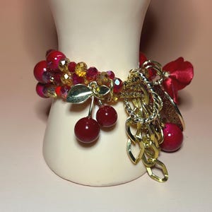 May include: A beaded bracelet featuring red and gold beads, with cherry charms and gold chain accents. The bracelet includes a red satin bow and gold leaf charms. The bracelet is displayed on a cream-colored form.