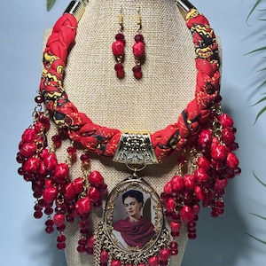 Könnte beinhalten: Eine rote Stoffkette mit einem Frida Kahlo-Anhänger und passenden Ohrringen. Die Halskette hat ein geflochtenes Design mit goldenen Akzenten und roten Perlenclustern. Der Anhänger ist ein ovales Porträt von Frida Kahlo.