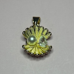 May include: A gold-toned pendant shaped like an open seashell, featuring a single round white pearl on the left side and a clear, pear-shaped faceted gemstone on the right side, complete with a top bail for attachment to a chain.