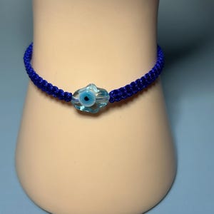 Könnte beinhalten: Ein blaues Makramee-Armband mit einem transparenten, hellblauen Glasanhänger mit einem blauen Augendesign. Das Armband wird auf einer hellen Form vor blauem Hintergrund präsentiert. Das gewebte Design des Armbands ist sichtbar.