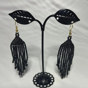 Puede incluir: Pendientes de flecos de cuentas negras con ganchos dorados. Los pendientes se exhiben en un soporte de metal negro con forma de hojas. Los pendientes tienen forma de lágrima.