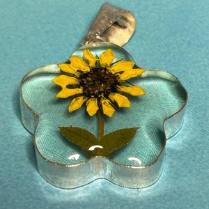 May include: A flower-shaped pendant with a real yellow sunflower encased in clear resin. The pendant has a silver-colored metal border and a bail for a necklace chain. The sunflower has a dark brown center and green leaves.