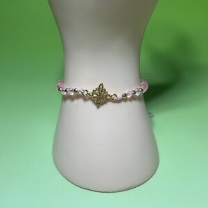 May include: A delicate pink beaded bracelet with a gold-colored Celtic knot charm. The bracelet features small, clear beads and gold accents, displayed on a white mannequin hand against a green background.