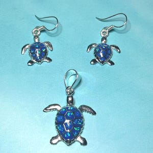 May include: A jewelry set featuring a turtle design. The set includes a pendant and a pair of earrings. Each piece has a silver-colored metal frame and a blue, mosaic-like center. The earrings have a hook closure.