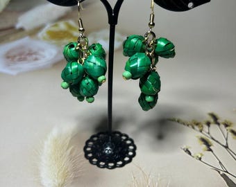 May include: A pair of green woven earrings with a cluster design, hanging from gold-colored hooks. The earrings are displayed on a black earring stand with leaf-shaped accents. The earrings have small green beads.