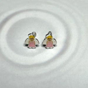 A pair of silver earrings with small, cartoon-like figures in pink and white colors, placed on a white surface with a wavy pattern.