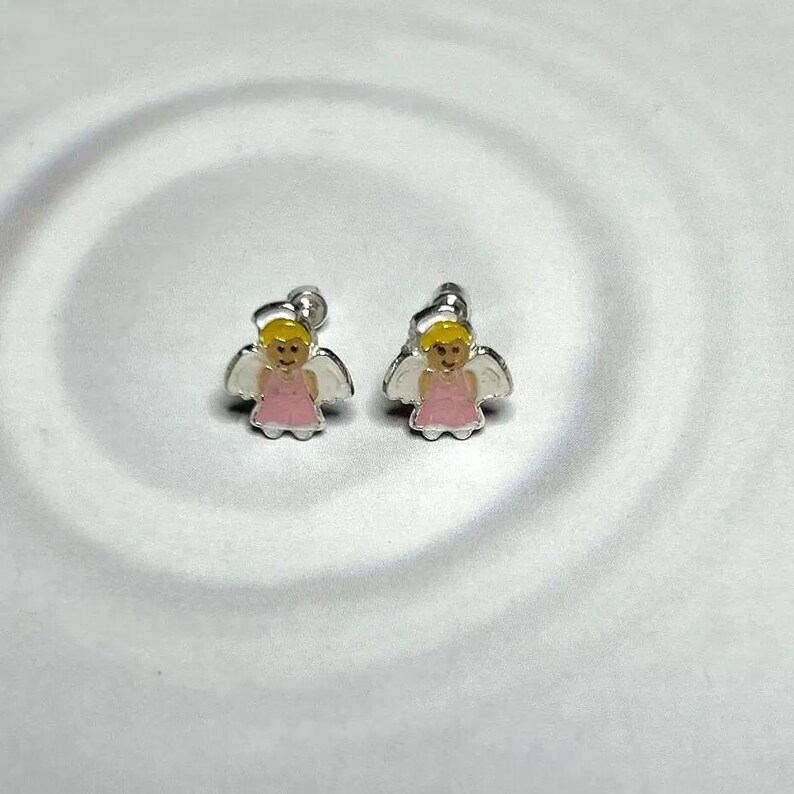 A pair of silver earrings with small, cartoon-like figures in pink and white colors, placed on a white surface with a wavy pattern.