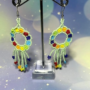 Puede incluir: Un par de pendientes de cuentas con un diseño circular y hebras colgantes. Los pendientes presentan una base verde claro con cuentas de colores rojo, amarillo, azul y verde. Los pendientes se exhiben en un soporte plateado para pendientes.