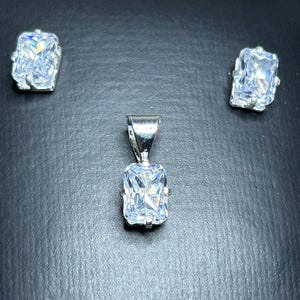 Puede incluir: Un colgante de plata con una gema rectangular y transparente, flanqueado por dos pendientes a juego. El conjunto de joyas se exhibe sobre una superficie oscura, resaltando el brillo de las piedras.