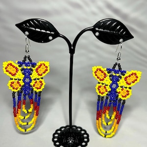 Puede incluir: Un par de pendientes de mariposa con cuentas. Los pendientes presentan cuentas amarillas, rojas y azules. Las alas de la mariposa son amarillas con detalles rojos, y el cuerpo es azul. Los pendientes cuelgan de ganchos plateados.