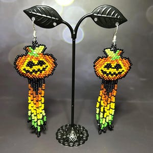 Puede incluir: Un par de pendientes de cuentas de Halloween con forma de calabazas. Los pendientes presentan cuentas naranjas, amarillas, verdes y negras, con un diseño de cara sonriente. Cuelgan de ganchos plateados.