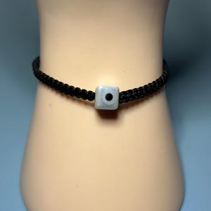 May include: A black braided cord bracelet with a square, light gray bead at the center. The bead has a small, dark dot in the middle. The bracelet is displayed on a pale, flesh-toned surface.