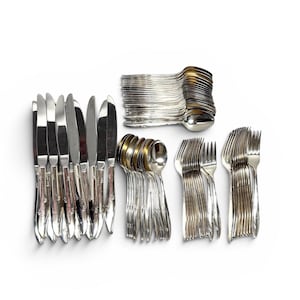 May include: A collection of silver-colored flatware, including knives, spoons, and forks, arranged on a white surface. The knives have a shiny, reflective blade. The spoons and forks are neatly stacked in separate piles.