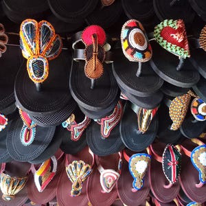 May include: A collection of beaded sandals in various colors and designs. The sandals feature intricate beadwork in patterns like hearts, circles, and abstract shapes. The soles are black or brown, and the beads are in shades of red, orange, blue, and white.