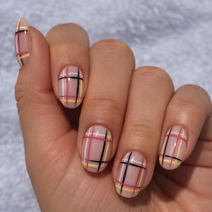 May include: A hand with oval-shaped nails painted with a plaid design. The nails are a nude color with a pattern of thin black, red, and yellow lines. The design is a grid pattern.