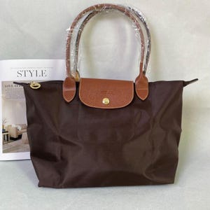 May include: A dark brown tote bag featuring a tan leather flap with a gold-tone snap closure. The bag has two long brown leather handles, one of which is wrapped in clear plastic. The zip pull is gold-tone and circular with an embossed logo.