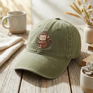 May include: Olive green baseball cap featuring an embroidered cartoon monkey with a serene expression. The cap has a curved brim and a classic six-panel design. The monkey is brown with a light brown face and is sitting cross-legged.