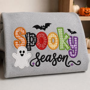 May include: A heather gray sweatshirt featuring embroidered text that reads Spooky season. The word Spooky is in colorful, patterned block letters with two black bats, and the word season is in black cursive with a white ghost icon.