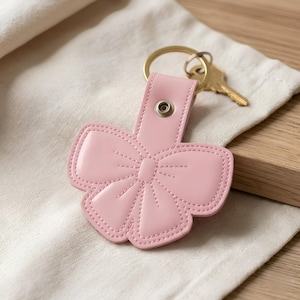 Può includere: Un portachiavi in pelle rosa chiaro a forma di fiocco con cuciture decorative. Il portachiavi presenta un bottone a pressione in metallo argentato sulla parte superiore ed è fissato a un anello in metallo dorato che regge una chiave color ottone.
