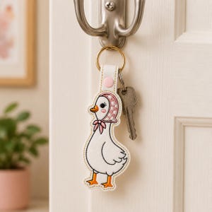 May include: A white duck-shaped keychain made of embroidered material, featuring a pink bonnet with white polka dots, orange feet, and a pink snap button. The keychain is attached to a gold-toned metal ring holding a single silver-colored metal key.