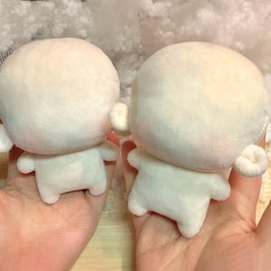 May include: Two plush dolls with rounded heads, small ears, and stubby arms and legs. The dolls are a soft, off-white color and are held in hands. The background is a light, fluffy material.