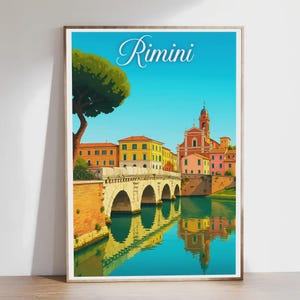 May include: A framed art print of Rimini, Italy, showing a bridge over a river with colorful buildings and a church. The sky is blue, and the water reflects the buildings. The word "Rimini" is written in cursive. The artwork has a bright, vibrant style.